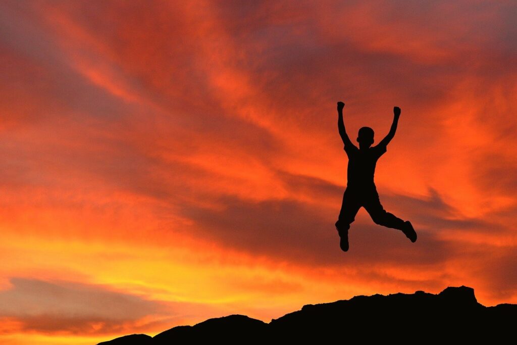 Silhouette of a boy leaping in the air with a dramatic orange clouds from a sunset with portions of a mountain in the background