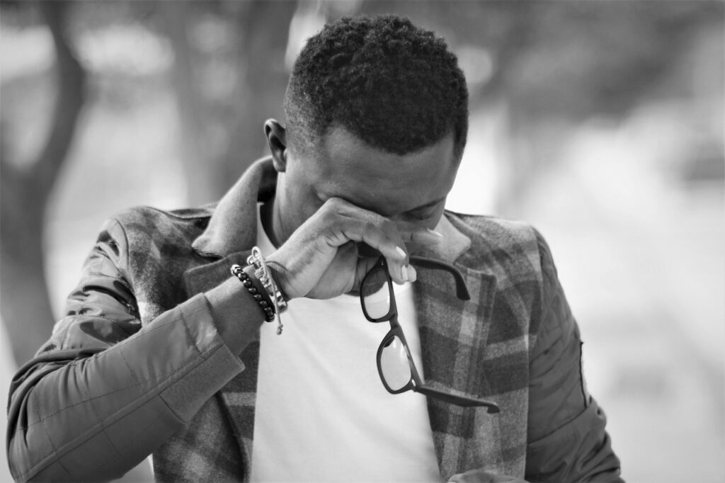 A black and white photo of an African man wearing a sport coat with head down and holding his right arm upward and wiping his eyes while holding a pair of glasses