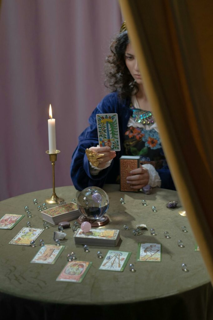 A female with black hair and light brown dress seated at a table filled with tarot cards, a candle, and a red rose while looking down at one of the cards held in her hands, and yet our future with God still is not known.