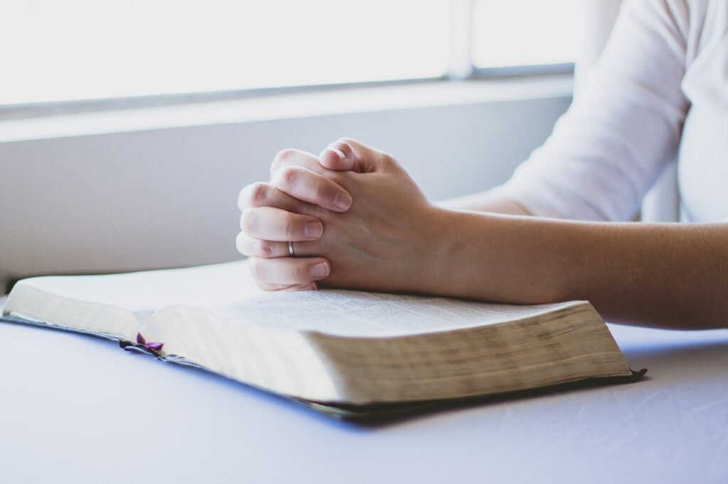 A person's hands clasped together and leaning on an open Bible symbolizing how God's victory comes through prayer, and trusting Him and His Word