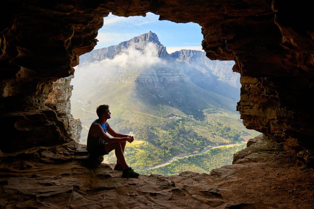 man sitting in opening of cave with sunlit mountain and valley in background symbolic of one contemplating on divine purpose