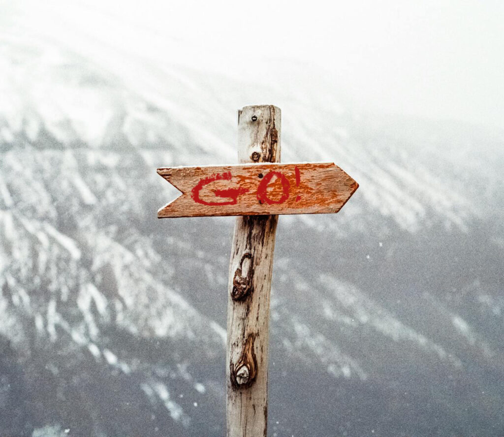 A rough wood post, set against a snowy mountainous background, with a wooden arrow attached pointing right with the word "GO!" written on it